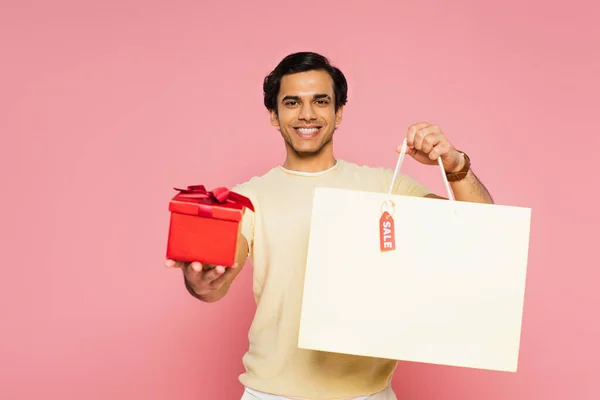 Heureux jeune homme tenant sac à provisions avec étiquette de vente et présent isolé sur rose — Photo de stock