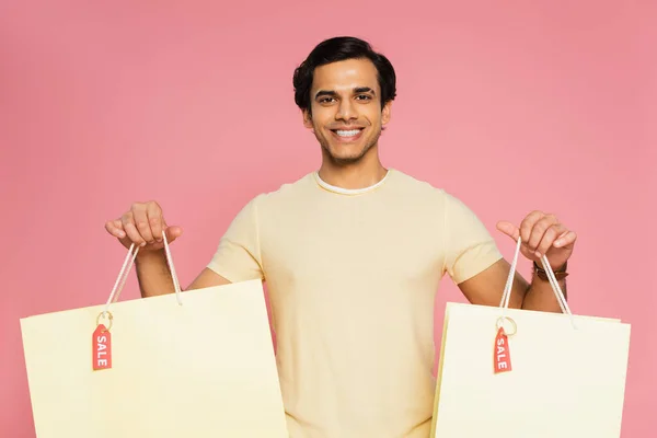Allegro giovane uomo che tiene borse della spesa con tag di vendita isolati su rosa — Foto stock
