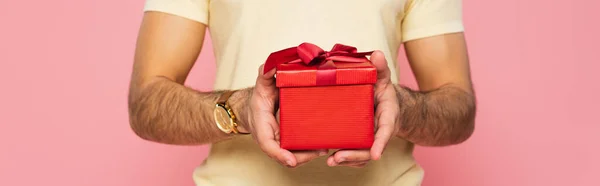 Vista recortada del hombre sosteniendo caja de regalo roja aislada en rosa, bandera - foto de stock