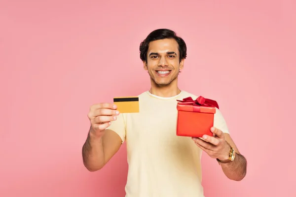 Joven alegre hombre sosteniendo caja de regalo roja y tarjeta de crédito aislado en rosa - foto de stock