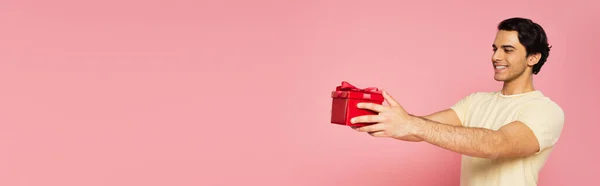 Joven hombre alegre sosteniendo caja de regalo roja aislada en rosa, bandera - foto de stock