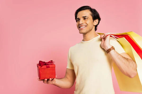 Smiling young man holding red gift box and shopping bags on pink — Stock Photo