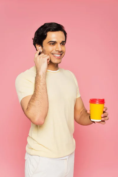 Усміхнений молодий чоловік тримає паперову чашку з кавою, щоб піти і налаштувати бездротові навушники ізольовані на рожевому — стокове фото