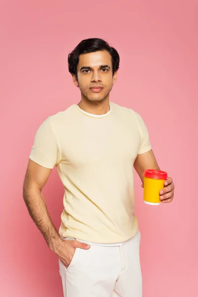 Young man holding paper cup with coffee to go while standing with hand in pocket isolated on pink — Stock Photo