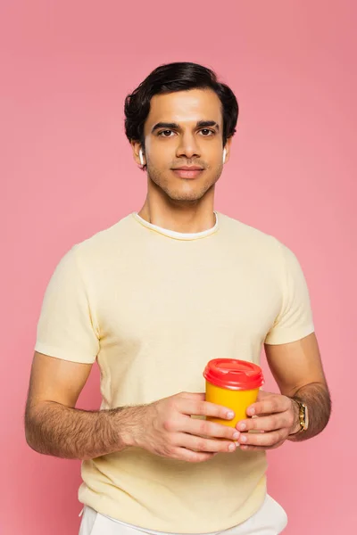 Heureux jeune homme dans les écouteurs sans fil tenant tasse en papier avec café pour aller isolé sur rose — Photo de stock