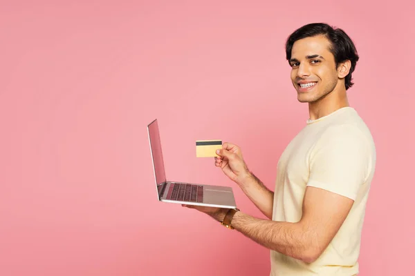 Young cheerful man holding credit card and laptop isolated on pink — Stock Photo