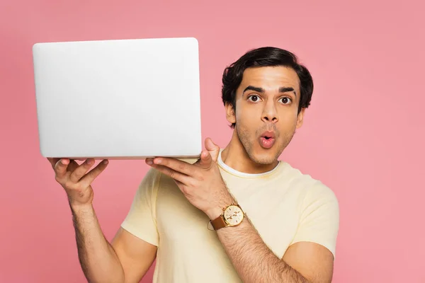Joven y sorprendido freelancer sosteniendo portátil aislado en rosa - foto de stock