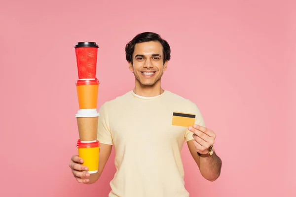 Joven feliz sosteniendo vasos de papel apilados y tarjeta de crédito aislada en rosa - foto de stock