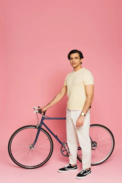 Молодой человек в полный рост стоит возле велосипеда на розовом — стоковое фото