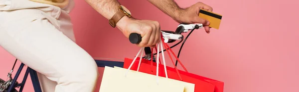 Partial view of young man riding bicycle with shopping bags and holding credit card on pink, banner — Stock Photo