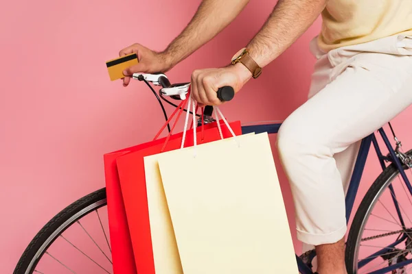 Partial view of young man riding bicycle with shopping bags and holding credit card on pink — Stock Photo