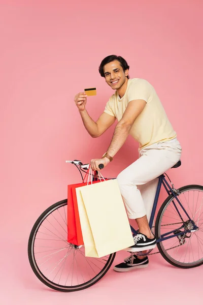 Heureux jeune homme à vélo avec des sacs à provisions et tenant carte de crédit sur rose — Photo de stock