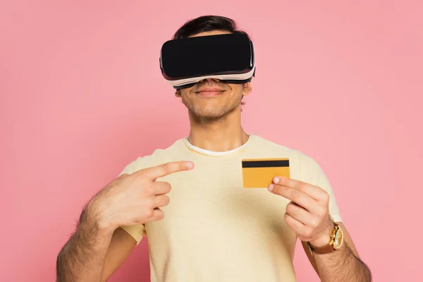 Веселий чоловік у гарнітурі vr, вказуючи пальцем на кредитну картку ізольовано на рожевому — стокове фото