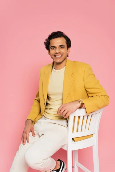 Cheerful young man in stylish blazer sitting on chair isolated on pink — Stock Photo