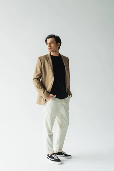 Voller Länge junger Mann in stilvollem beigen Blazer stehend mit Händen in Taschen auf grau — Stockfoto