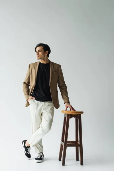 Young man in stylish beige blazer posing with hand in pocket near wooden chair on grey — Stock Photo