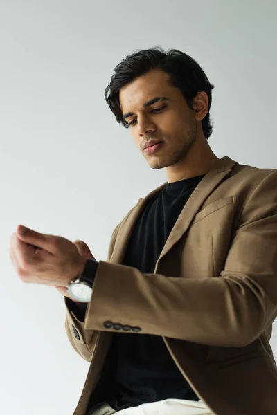 Young man in stylish beige blazer adjusting wristwatch isolated on grey — Stock Photo