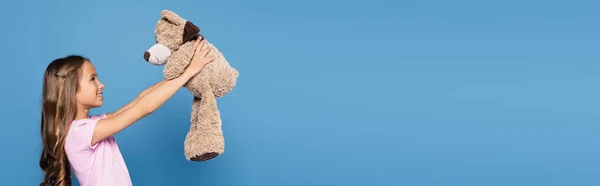 Vista laterale del bambino allegro che guarda il peluche isolato sul blu, banner — Foto stock