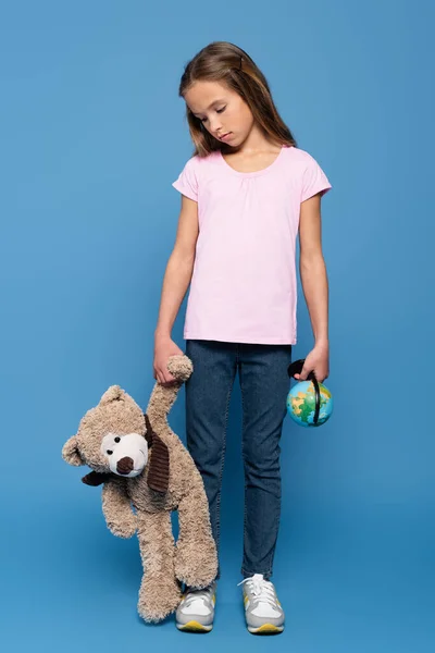 Trauriges Kind mit Globus und Teddybär auf blauem Hintergrund — Stockfoto