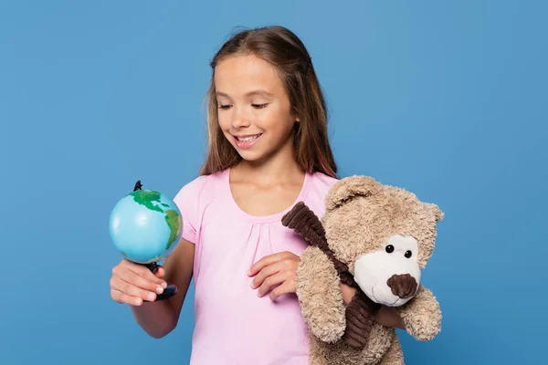 Preteen girl with globe and teddy bear smiling isolated on blue — Stock Photo