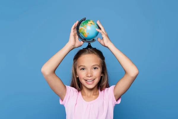 Kind lächelt in die Kamera und hält Globus auf dem Kopf isoliert auf blau — Stockfoto