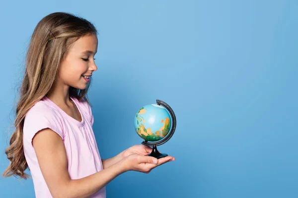 Side view of smiling girl holding globe on hands isolated on blue - foto de stock