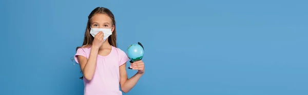 Écolier portant un masque médical et tenant le globe isolé sur bleu, bannière — Photo de stock