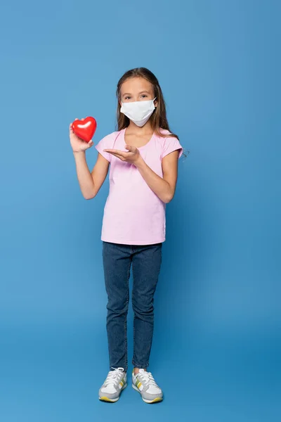 Girl in protective mask holding red decorative heart and pointing with hand on blue background - foto de stock