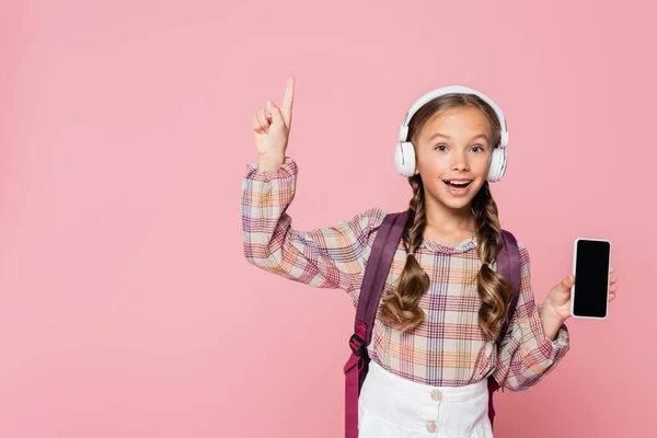 Positives Schulkind mit Kopfhörer hat Idee, während es Smartphone isoliert auf rosa hält — Stockfoto