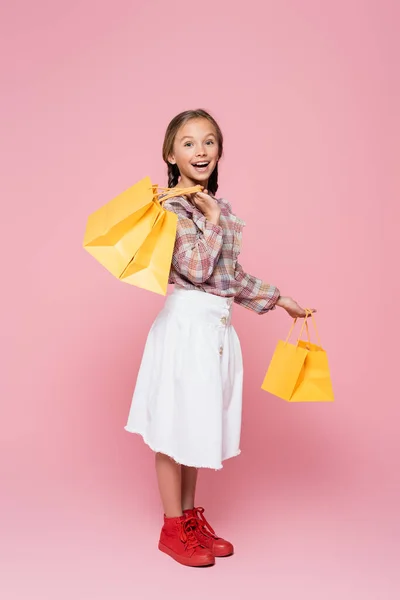 Amazed kid in plaid blouse and white skirt holding yellow shopping bags on pink background — Photo de stock