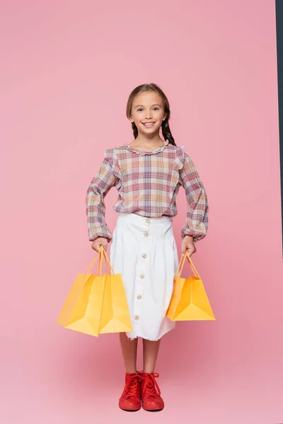 Повний вигляд щасливої дівчини з жовтими торговими пакетами на рожевому фоні — стокове фото