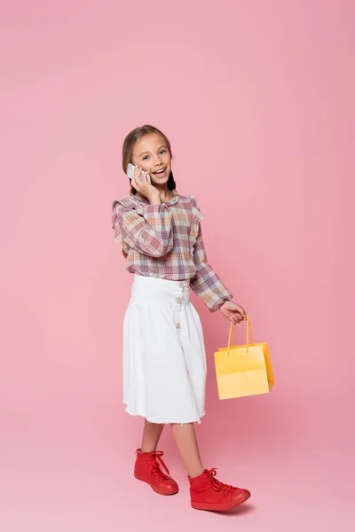 Criança na moda falando no smartphone enquanto caminha com saco de compras amarelo no fundo rosa — Fotografia de Stock