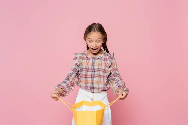Bambino sorpreso in camicetta a quadri guardando nella shopping bag gialla isolato su rosa — Foto stock