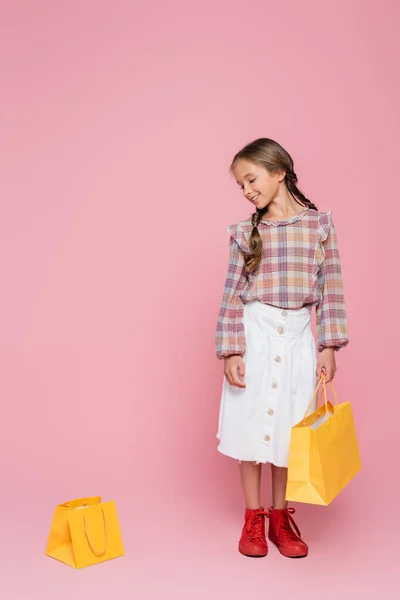 Smiling girl in white skirt and plaid blouse looking at yellow shopping bag on pink background - foto de stock