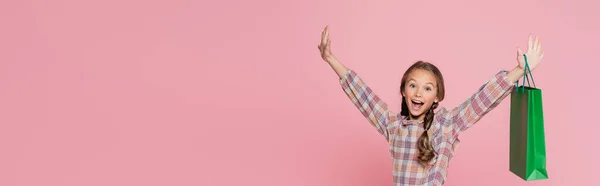 Erstauntes Mädchen mit grüner Einkaufstasche und erhobenen Händen, Kamera isoliert auf rosa, Banner — Stockfoto
