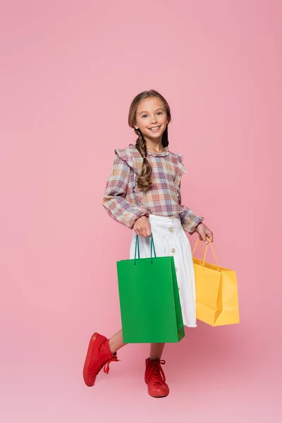 Smiling child in stylish clothes holding green and yellow shopping bags on pink background — Stock Photo