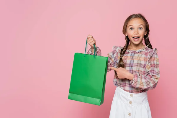 Erstauntes und glückliches Kind zeigt auf grüne Einkaufstasche auf rosa — Stockfoto