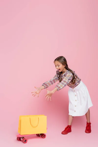 Excited girl with open mouth and outstretched hands near yellow shopping bag and penny board on pink background — Stock Photo