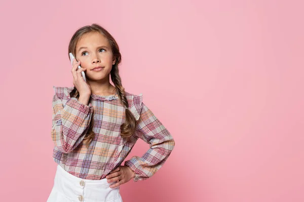 Serious preteen girl talking on smartphone while standing with hand on hip isolated on pink - foto de stock