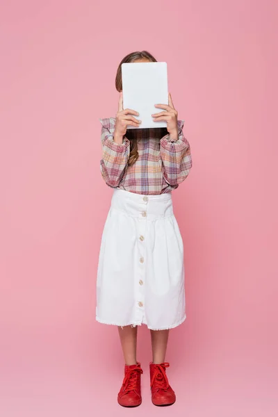 Girl in white skirt, plaid blouse and red sneakers obscuring face with digital tablet on pink background - foto de stock