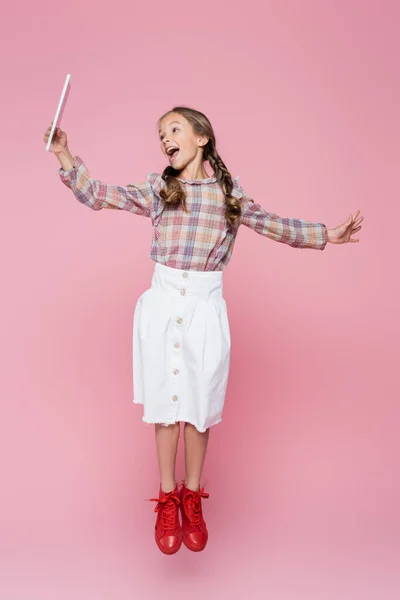 Изумленный ребенок в модной одежде левитации с цифровой таблеткой на розовом фоне — стоковое фото
