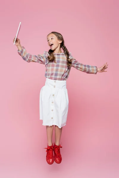 Astonished child in trendy clothes levitating with digital tablet on pink background — Stock Photo