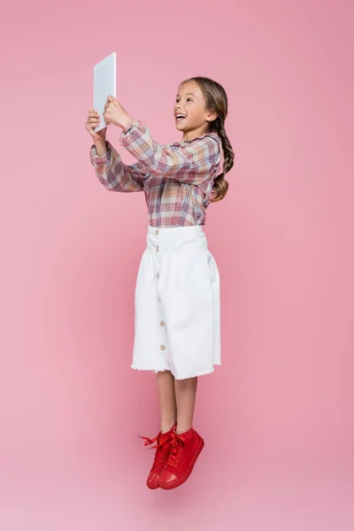 Aufgeregtes Mädchen in stilvoller Kleidung schwebt mit digitalem Tablet auf rosa Hintergrund — Stockfoto