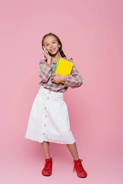 Visão de comprimento total da moda, menina alegre com livros falando no celular no fundo rosa — Fotografia de Stock