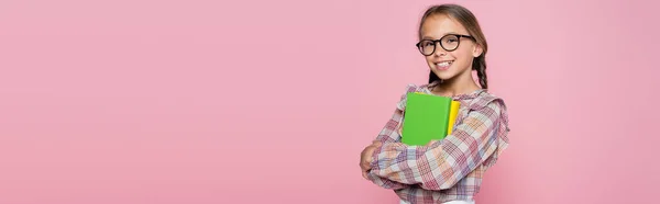 Ragazza sorridente in camicetta a quadri e occhiali in piedi con libri isolati su rosa, striscione — Foto stock