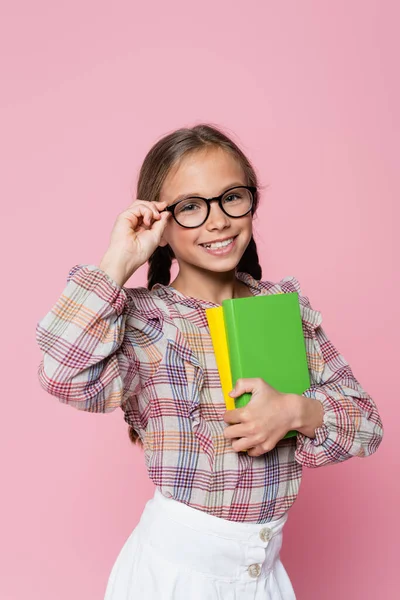 Positives Mädchen berührt Brille, während es Lehrbücher isoliert auf rosa hält — Stockfoto