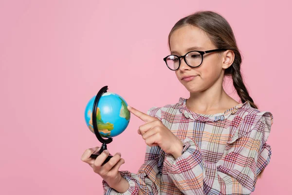 Scettico ragazza in occhiali che punta a piccolo globo isolato su rosa — Foto stock