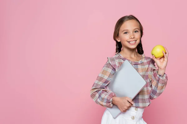 Preteen ragazza con mela fresca e laptop sorridente alla fotocamera isolata su rosa — Foto stock