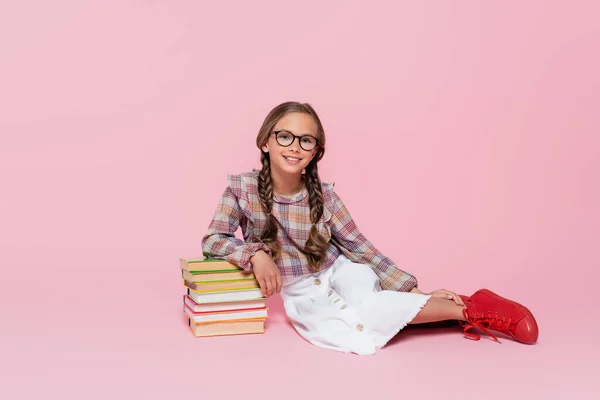 Lächelndes Kind in stylischer Freizeitkleidung, das neben einem Stapel Bücher auf rosa Hintergrund sitzt — Stockfoto