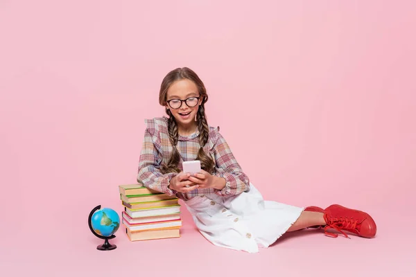 Glückliches Mädchen, das neben Bücherstapel und Globus sitzt, während es auf dem Handy auf rosa Hintergrund plaudert — Stockfoto